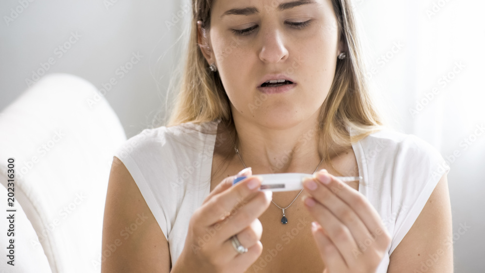 Photo of young sick woman sitting on sofa and holding thermometer Photo of young sick woman sitting on sofa and holding thermometer