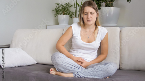 Young woman in pajamas sitting on sofa and suffering from menstrual pain