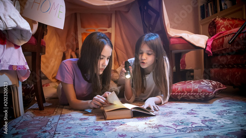 Two sisters in pajamas lying on floor at selfmade tent and reading book with flashlight