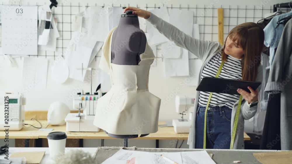 Beautiful tailor is turning tailoring dummy to check pinned clothing and working with tablet to enter information. Using modern technologies in clothes manufacturing concept.