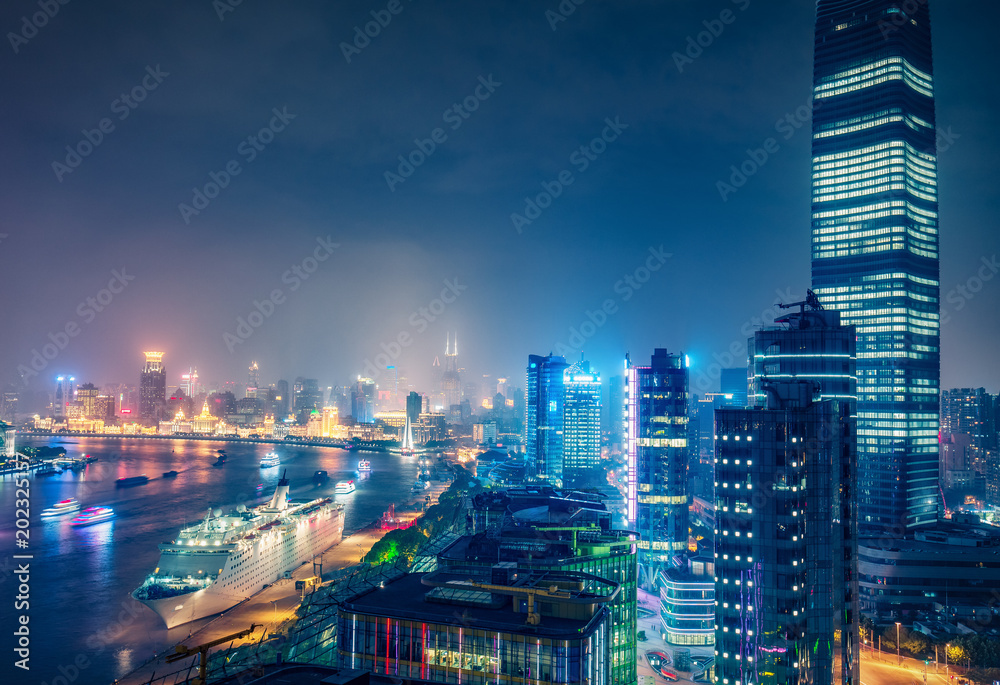 Fototapeta premium Nocna panorama Szanghaju w Chinach. Spektakularny widok z lotu ptaka na oświetlone wieżowce i rzekę Huanpu z łodziami.