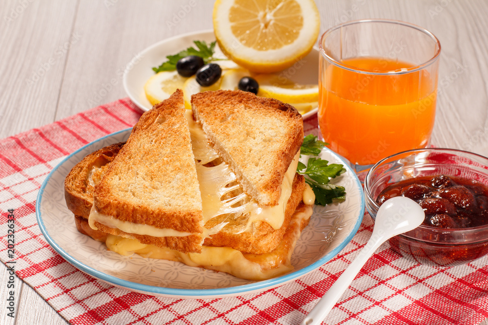 Toasted slices of bread with cheese and green parsley on white plate, glass of orange juice, lemon, olives, spoon and bowl with strawberry jam on napkin