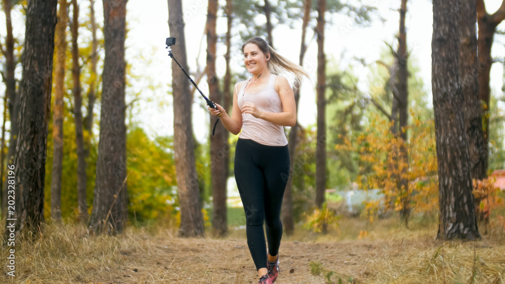 Young sporty woman running with camera on selfie stick at forest
