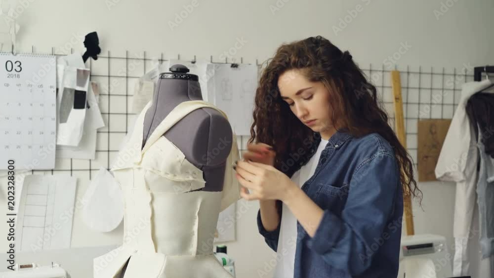 Young pretty seamstress is busy adjusting clothing on tailoring dummy with sewing pins. Women's garments, sketches on wall, tailoring tools are visible. Young pretty seamstress is busy adjusting clothing on tailoring dummy with sewing pins. Women's garments, sketches on wall, tailoring tools are visible.