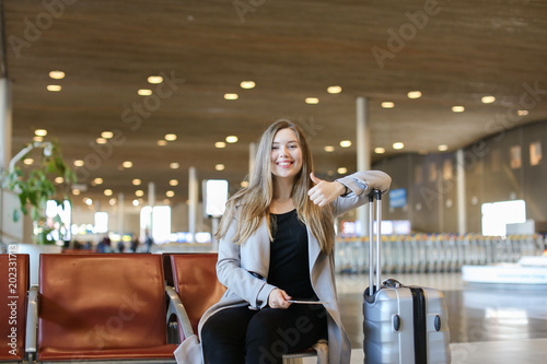Nice girl making video call by modern tablet in airport waiting room and sitting near grey valise, showing thumbs up. Concept of communication, free hotspot and traveling abroad.