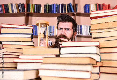Time flow concept. Man, scientist looks at hourglass. Teacher or student with beard studying in library. Man on serious and shocked face watching time is going over, bookshelves on background.