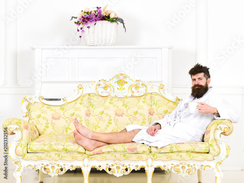 Man sleepy in bathrobe drinks coffee in luxury hotel in morning, white background. Man with beard and mustache enjoys morning while sitting on old fashioned luxury sofa. Luxury life concept. photo