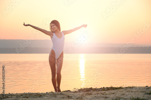 Young woman in strings swimsuit stands on the beach in sunrise