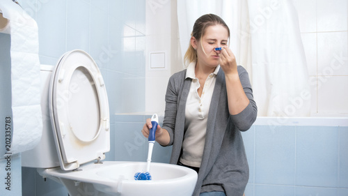 Young woman cleaning cleaning toilet feeling bad smell and closing nose with clothespin photo