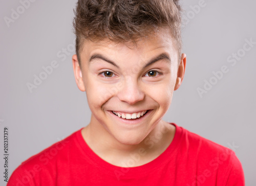 Silly teen boy making grimace - funny face. Child on gray background. Emotional portrait of caucasian teenager looking at camera.