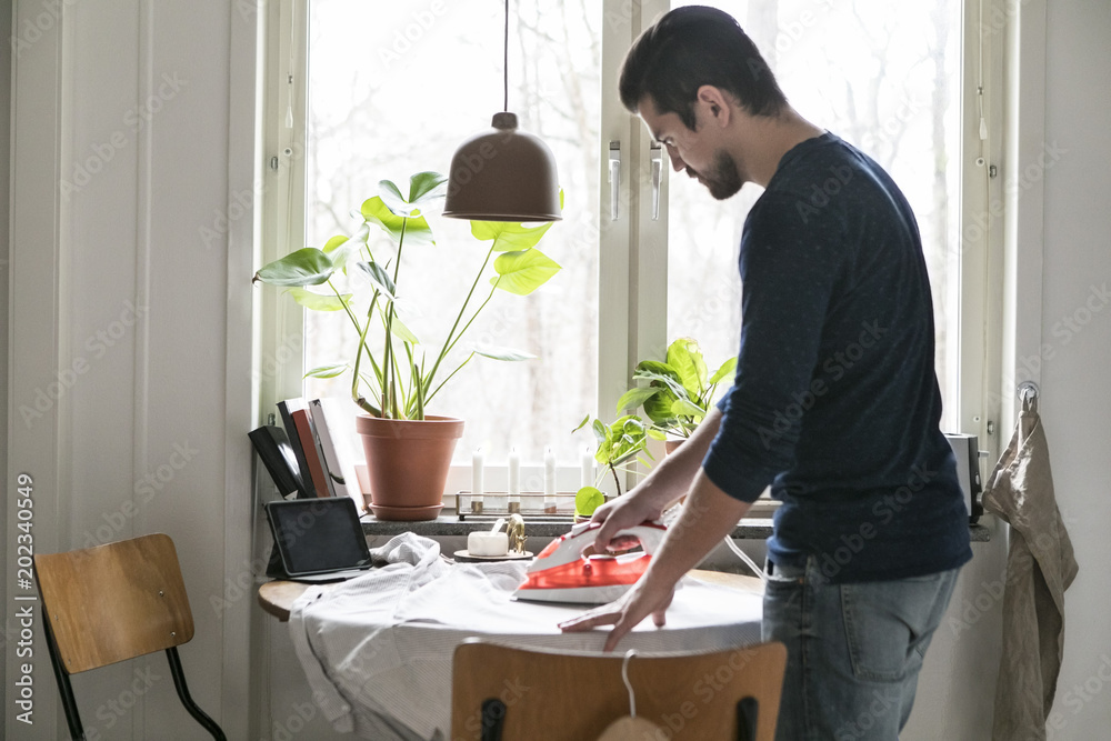 Side view of man ironing shirt on table by window while standing at kitchen Side view of man ironing shirt on table by window while standing at kitchen