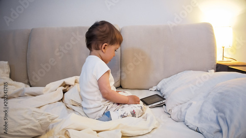 Adorable toddler boy in pajamas sitting o nbed at night and watching cartoons on digital tablet photo