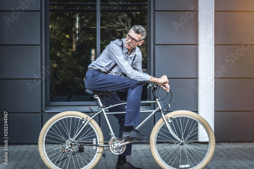 Fototapeta Naklejka Na Ścianę i Meble -  Modern lifestile - modern business. Side view of mature businessman looking over shoulder while riding on his bicycle.