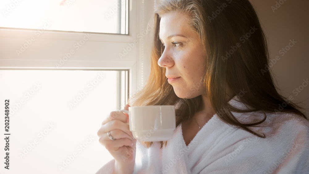 Beautiful brunette woman in bathrobe looking at sunrise out of window and drinking coffee Beautiful brunette woman in bathrobe looking at sunrise out of window and drinking coffee