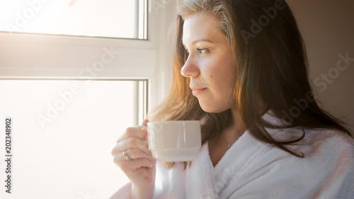 Beautiful brunette woman in bathrobe looking at sunrise out of window and drinking coffee