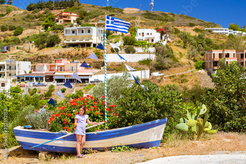 Girl with yellow lemon in hand posing against old fishing boat with Greek flag, stands on shore and decorated by flower bed. Tourist attraction Varkotopos beach in resort village Bali, Crete, Greece photo