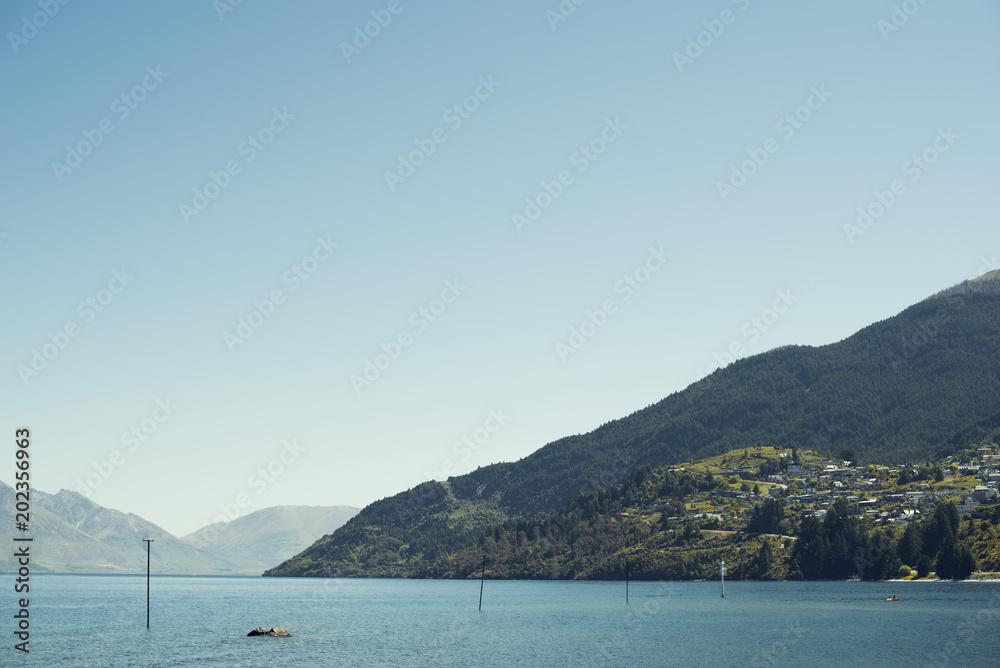 Paisaje de montaña verde bajo cielo azul despejado frente un lago en Nueva Zelanda.	