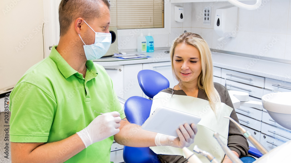 Portrait of male dentist explaining teeth treatment to young woman using digital tablet