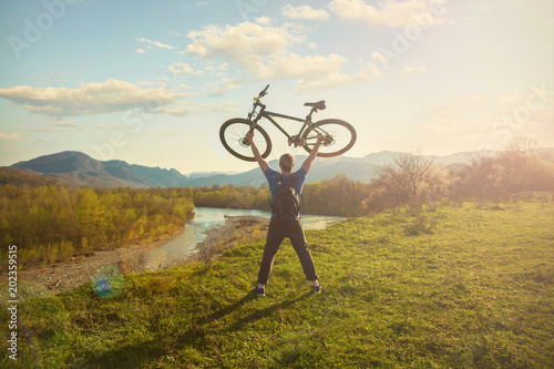 Boy cyclist standing on a mountain and looks at the river at sunset A cyclist holds a bicycle over his head.
