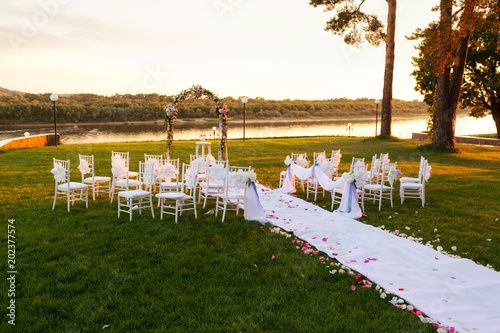 sunset on river. Romantic wedding ceremony. White wooden chairs with ribbon and flowers on a green lawn. Nice armchairs for guests