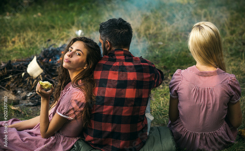 Brunette girl leaning on her boyfriend. Turn back friends sitting on grass next to campfire. Romantic weekend in nature