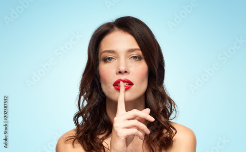 beauty, secret, make up and people concept - woman holding finger on lips or mouth with red lipstick over blue background
