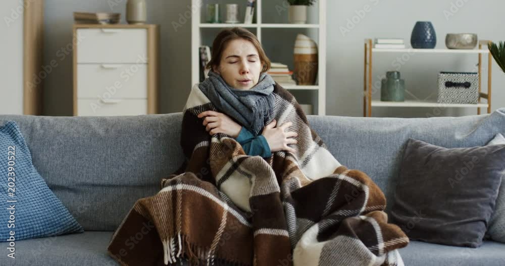 Caucasian young ill woman in the warm scarf shivering under the plaid on the couch in the living room as she having a high temperature during a flu. At home. Indoor