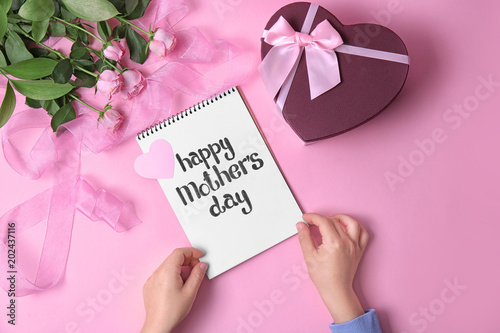 Child holding notebook with phrase "HAPPY MOTHER'S DAY" near gift and roses on table
