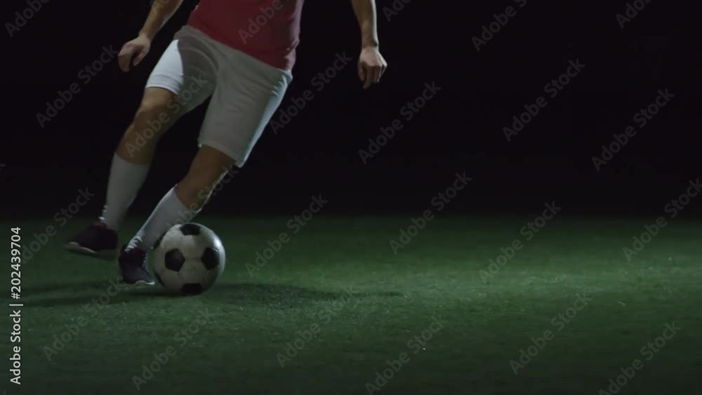 Tilt up of young female soccer player dribbling a ball on field with artificial turf in dark indoor arena Tilt up of young female soccer player dribbling a ball on field with artificial turf in dark indoor arena