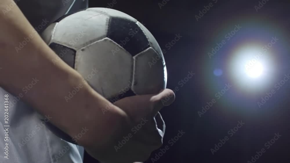 Closeup of professional player holding soccer ball and walking on stadium in darkness with projector lightning in background