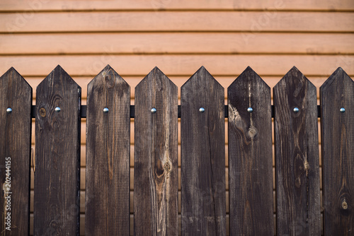 Pointed wooden fence on backdrop of house with surface from siding. Background image of wooden fence from rectangular planks with triangular top. Planks fastened crossbar and nails.