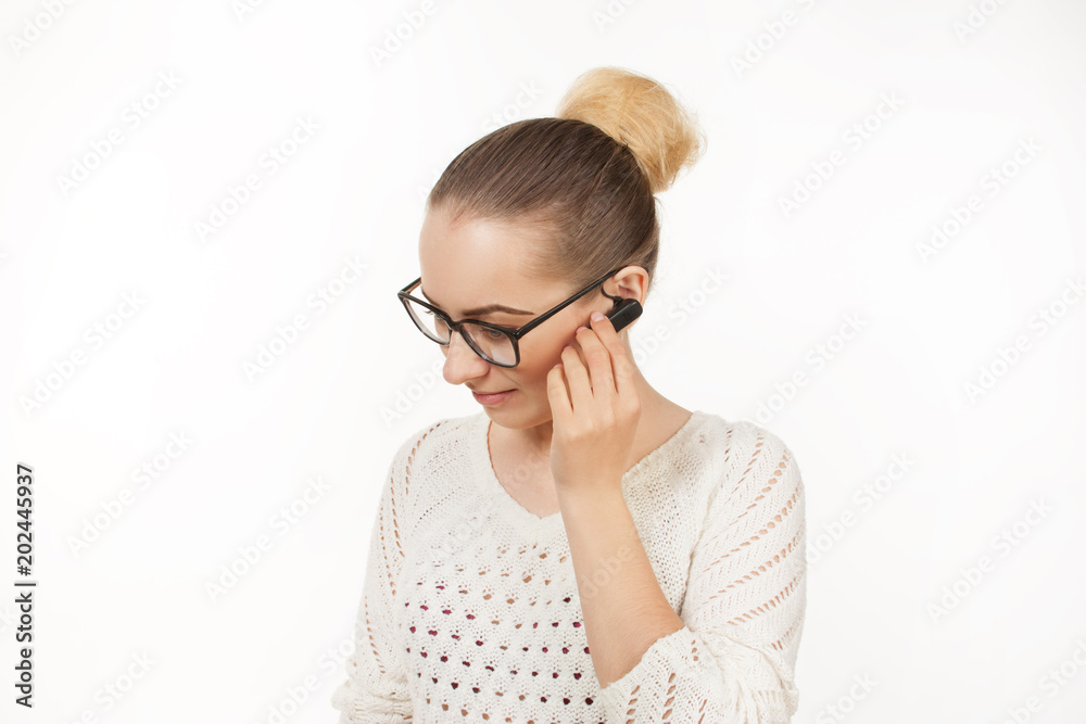 beautiful blonde girl speaks on the phone through a bluetooth earpiece on a white background. emotionally speaking girl in glasses with gathered hair speaks on the phone