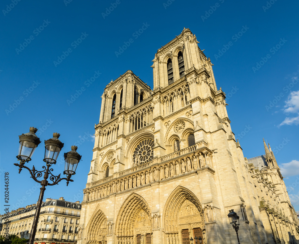 View of the facade and bell towers of Notre-Dame de Paris cathedral, showing the three portals and the rose window in the warm light of the setting sun, with a vintage street light in the foreground.