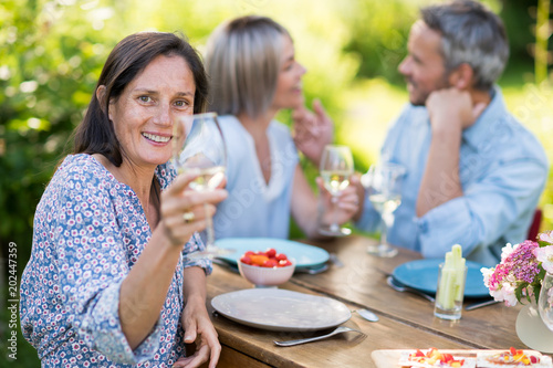 In summer. a group of friends in their forties gathered around a table in the garden to share a meal. They toast with their glasses of wine to the camera.
