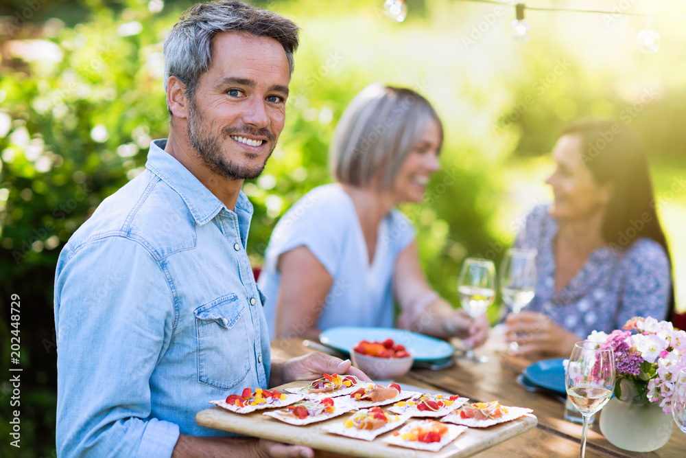 group of friends in their forties gathered around a table in the garden to share a meal. A man offers snacks to guests while looking at the camera group of friends in their forties gathered around a table in the garden to share a meal. A man offers snacks to guests while looking at the camera