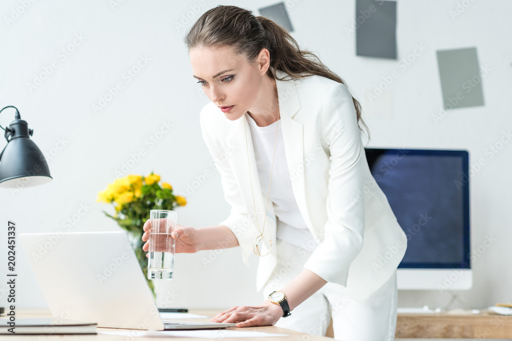 focused businesswoman with glass of water using laptop at workplace in office focused businesswoman with glass of water using laptop at workplace in office