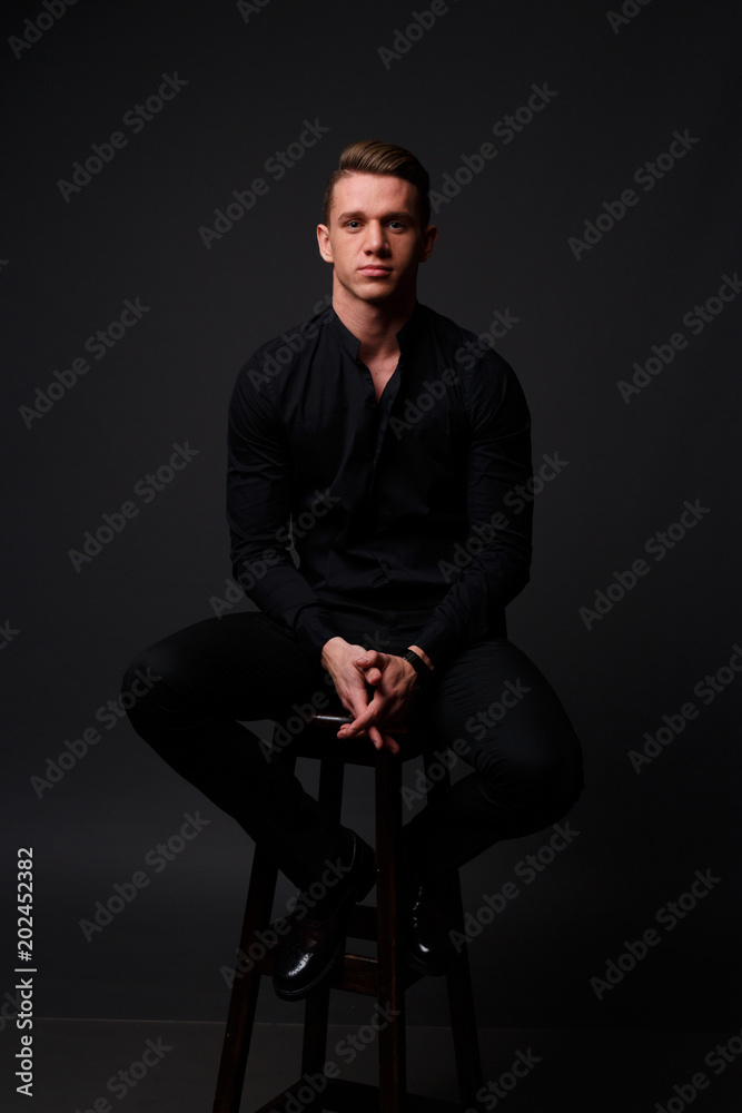a young, white guy sits on a chair in a black shirt and black pants,  against a black background and looking at camera. business, model,  interview, portrait Stock Photo
