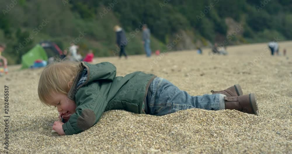 A little toddler boy is lying face down on the beach and is playing with pebbles. Slow motion handheld shot. A little toddler boy is lying face down on the beach and is playing with pebbles. Slow motion handheld shot.