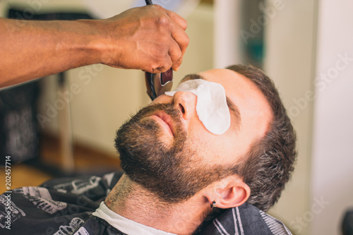 Close up view of man with eye protectors at barbershop. Barber cutting the beard with the trimmer.