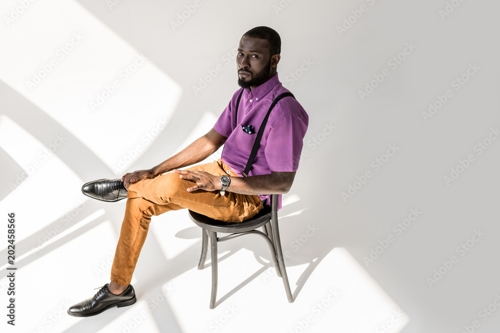 side view of thoughtful african american man in fashionable clothing sitting on chair on grey background