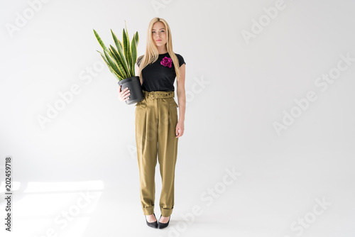 full length view of stylish blonde girl holding potted plant and looking at camera on grey