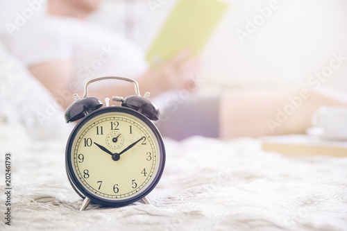 Young man resting on a beed reading a book. Alarm clock and empty copy space for Editor's text.