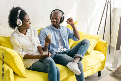 cheerful african american listening music in headphones with smartphones while resting on sofa at home photo