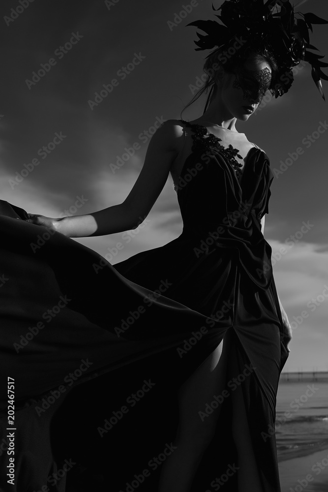 Black and white low key portrait of beautiful young woman in chic black dress, big floral crown and lace mask on face. Sea on background
