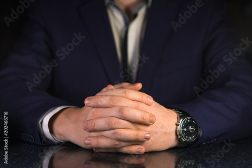 Businessman in blue suit and white shirt with crossed fingers sitting by the table on dark background