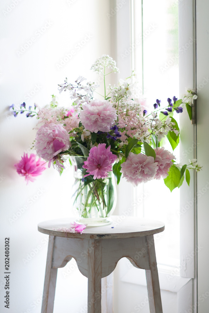 A vase of colorful pink and purple flowers sits on a white pedestal next to a window and door. The flowers are fresh, blooming, large, and beautiful.
