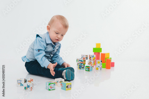 blond boy in blue suit plays with colorful blocks on white background. A small child builds a tower of cubes on the floor