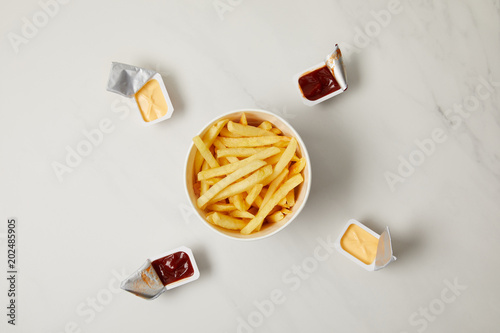 top view of tasty french fries in bowl surrounded with containers of sauces on white