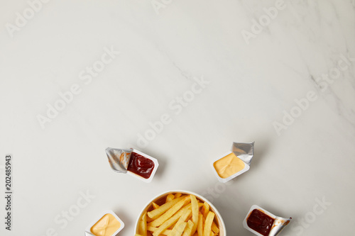 top view of french fries in bowl surrounded with containers of sauces on white