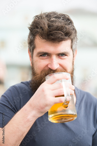 Bearded man holds beer mug, drinks beer outdoor. Craft beer concept. Man with long beard looks relaxed. Man with beard and mustache on happy smiling face, light background, defocused.